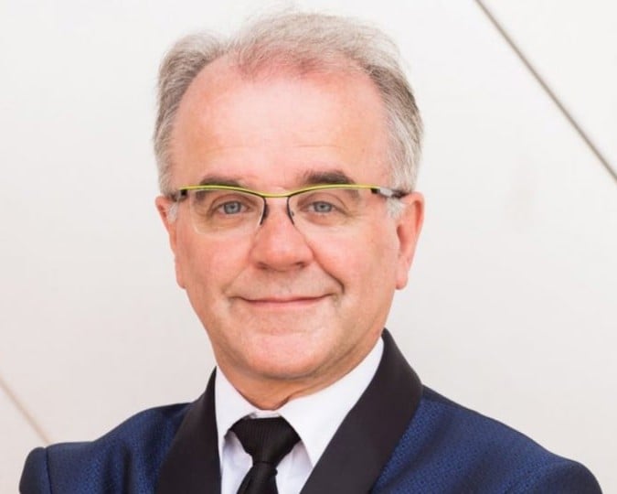 Man in patterned blue dinner jacket , black tie, and vivid yellow glasses poses.