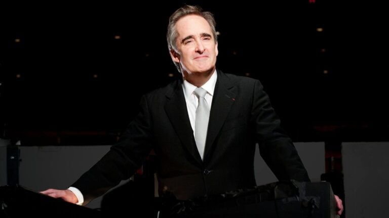 James Conlon wearing a dark suit, white shirt, light grey tie, posing in the orchestra pit of LA Opera's Dorothy Chandler Pavilion.