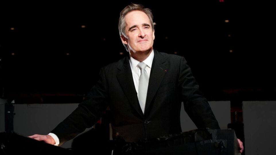 James Conlon wearing a dark suit, white shirt, light grey tie, posing in the orchestra pit of LA Opera's Dorothy Chandler Pavilion.