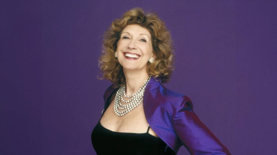 Felicity Lott smiling widely, in a sleeveless black gown with a purple bolero jacket and a layered rhinestone necklace; purple backdrop mathches color of jacket