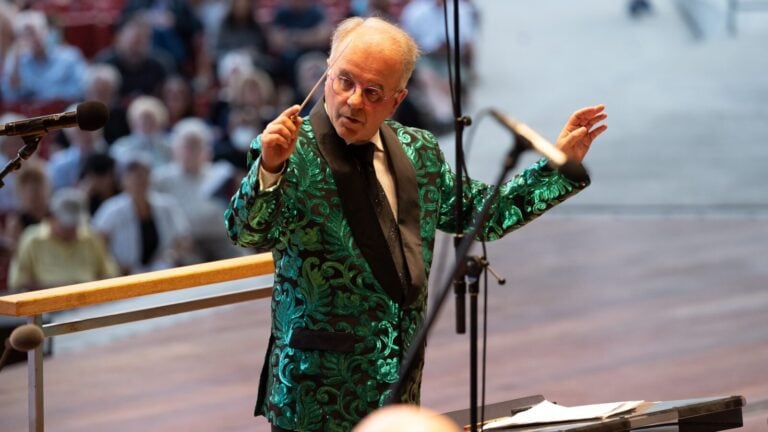 Christopher Bell in an ornate green tuxedo jacket conducting (musicians out of frame) at the Pritzker Pavillion, audience out of focus in background.