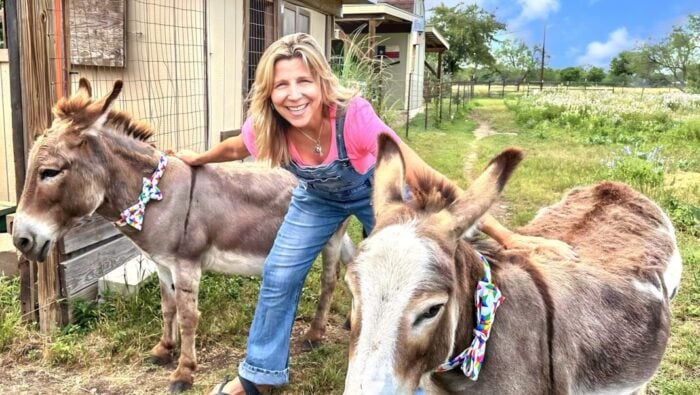 smiling woman in overalls flanked by 2 donkeys