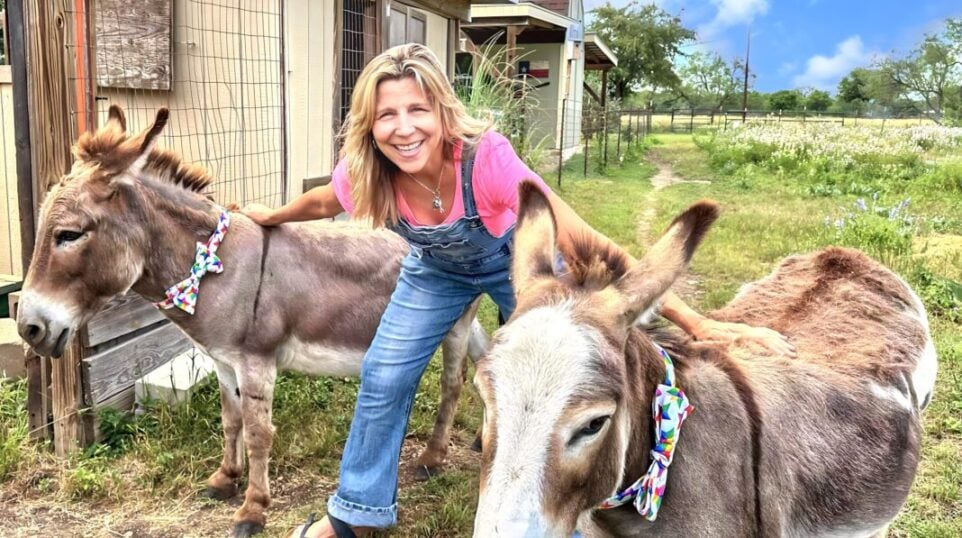 smiling woman in overalls flanked by 2 donkeys