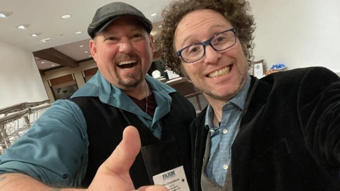 man in snap brim hat gives thumbs up next to a smiling man in glasses