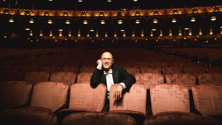 Enrique Mazzola in black tuxedo, white tie; sititng in the Ardis Krainik auditorium, lit from above, right hand resting on face.