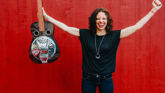 woman with Dobro in right hand raises arms in triumph