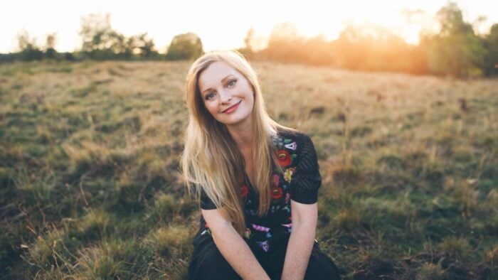 Lucy Crowe posing on a grassy bank, sun setting behind her which casts a light on her blonde hair softening the contrast of the portrait.