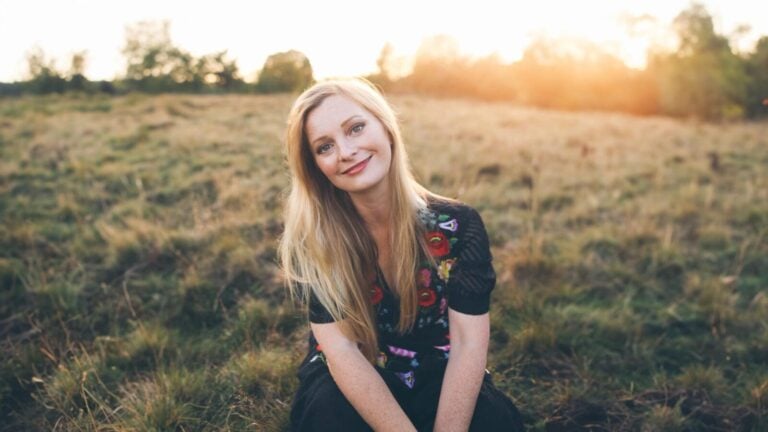 Lucy Crowe posing on a grassy bank, sun setting behind her which casts a light on her blonde hair softening the contrast of the portrait.