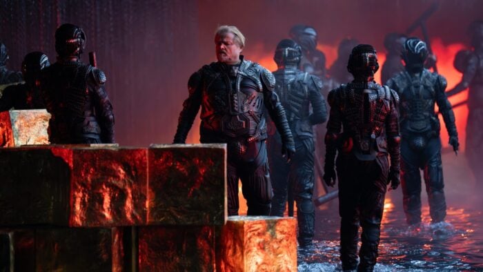 A middle aged white man with a short blond beard, clad in dark armor, looks at glowing pieces of magma stacked on top of each other like cubes.
