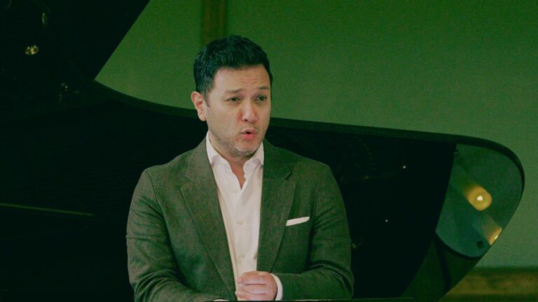 Performance video still of Nicholas Phan in light brown suit, white shirt, no tie; singing in the crook of a piano, lid on full stick.
