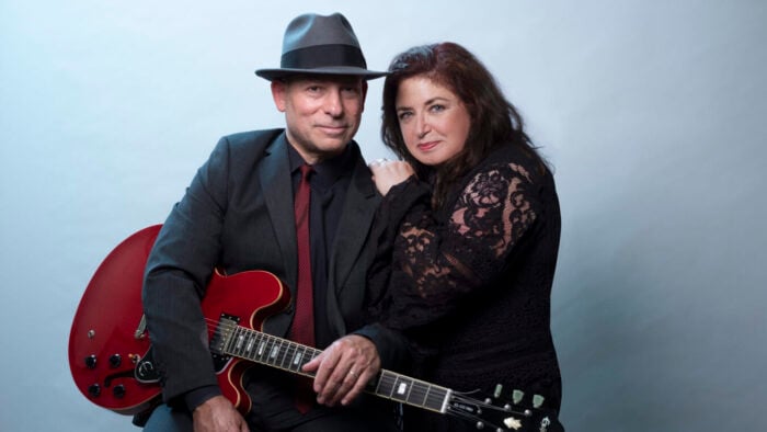 man with guitar wearing fedora, woman leans on his shoulder, both look into camera