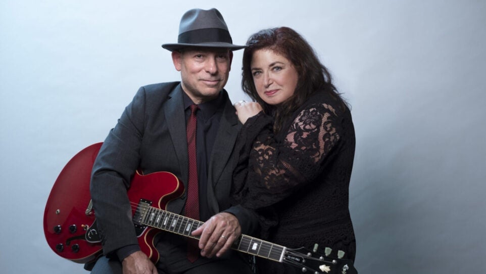 man with guitar wearing fedora, woman leans on his shoulder, both look into camera